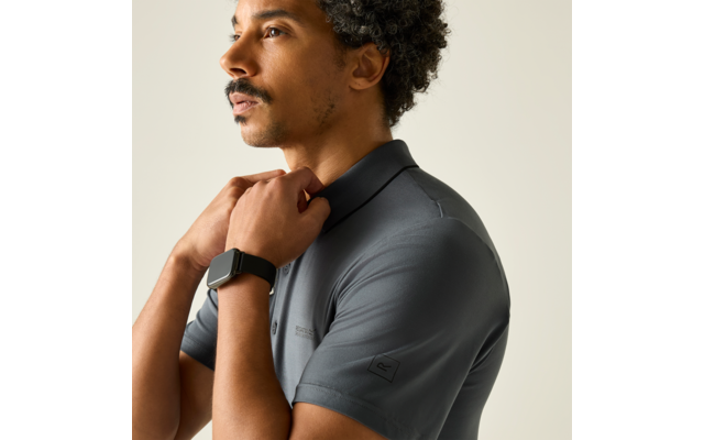 Mann im Profil, Schultern und Kopf sichtbar, lockige Haare, Schnurrbart, graues kurzärmeliges Poloshirt, Smartwatch am Handgelenk, Hände am Kragen, einfarbiger beiger Hintergrund