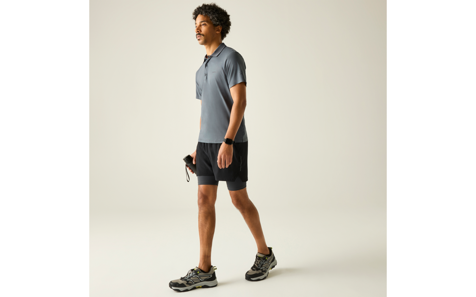 Seitliche Ganzkörperaufnahme eines Mannes in grauem Polo, schwarzen Shorts und Sportschuhen; Smartwatch am linken Handgelenk; er hält in der rechten Hand ein Springseil mit roten Griffen; einfarbiger heller Hintergrund