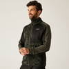 Homme aux cheveux foncés et barbe, debout devant un fond clair, porte une veste polaire zippée vert-noir avec fermeture éclair et petits logos sur la poitrine et la manche, regarde sur le côté, mains devant le corps