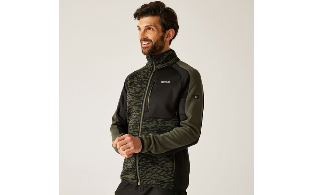 Homme aux cheveux foncés et barbe, debout devant un fond clair, porte une veste polaire zippée vert-noir avec fermeture éclair et petits logos sur la poitrine et la manche, regarde sur le côté, mains devant le corps