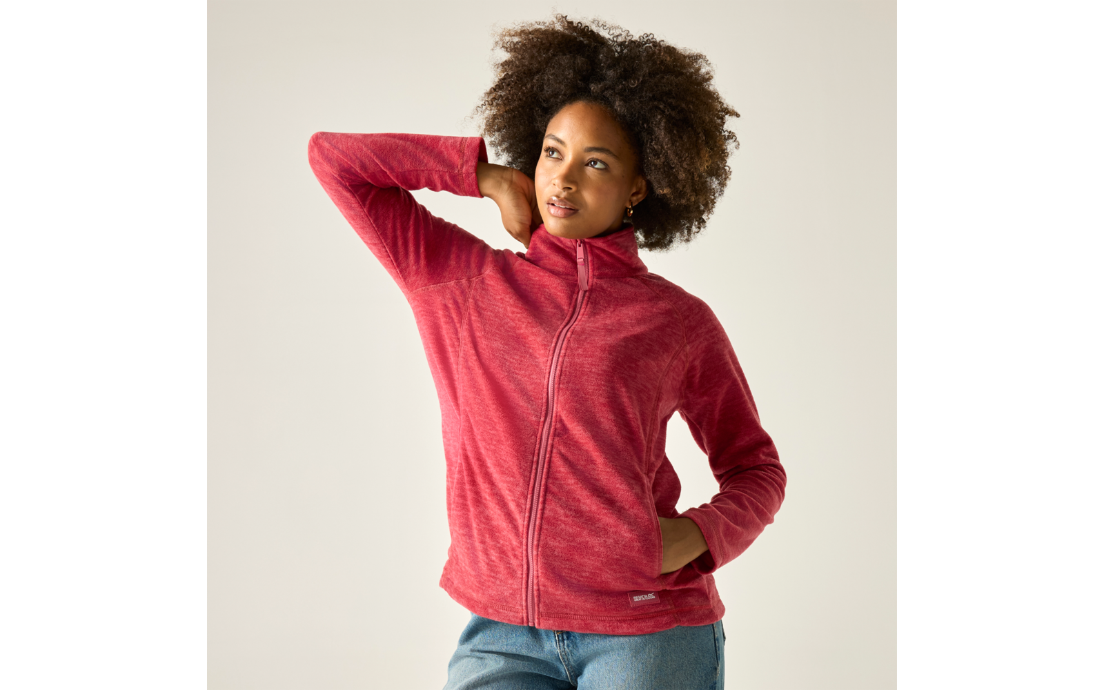 Person mit lockigem Haar trägt rote Fleecejacke mit Reißverschluss und Jeans, rechte Hand in Tasche, linke Hand hinter dem Kopf, heller Hintergrund, Blick zur Seite