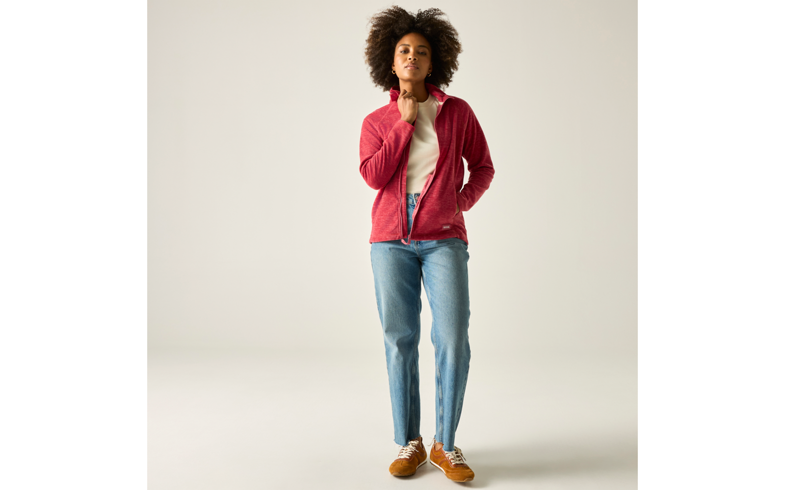 Person mit lockigem Haar steht; rote Jacke, weißes Shirt, blaue Jeans, braune Schuhe; rechte Hand am Hals, linke Hand in Tasche