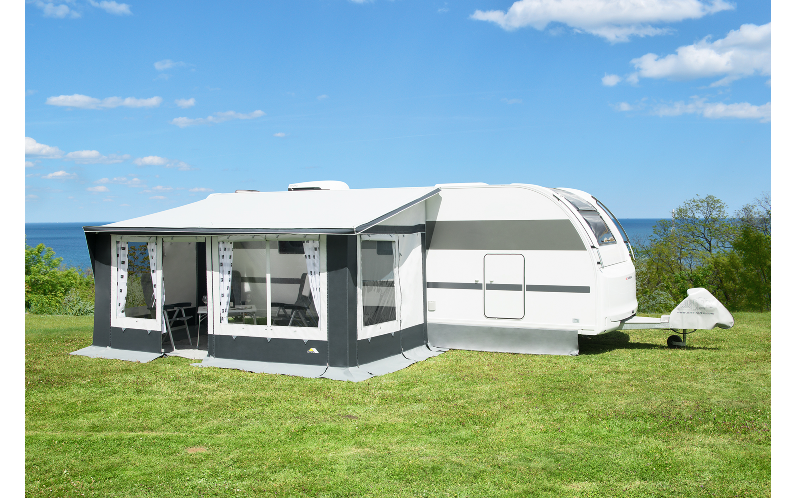 Weißer Wohnwagen mit grauem Vorzelt auf grünem Rasen, Tisch und Stühle im Vorzelt sichtbar, Meer im Hintergrund und blauer Himmel mit Wolken