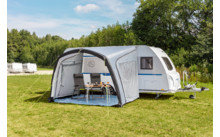 Witte caravan met grijs voortent; in het voortent een tafel en twee stoelen; grasveld, bomen en blauwe lucht met wolken