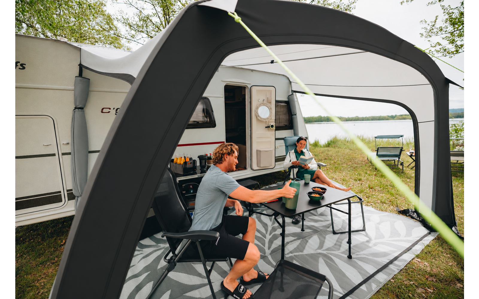 Due persone sono sedute a un tavolo pieghevole sotto una tenda gonfiabile accanto a una roulotte bianca; tazze e ciotole sono sul tavolo; sullo sfondo si vede un lago