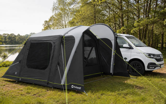 Graues Vorzelt an einem weißen Campingbus auf Gras, offener Eingangstunnel, grüne Abspannseile, See und Bäume im Hintergrund