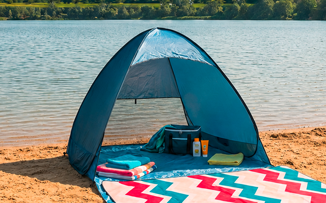 Tienda de playa azul en la orilla arenosa de un lago, manta con zigzag colorido delante, una bolsa y una botella de agua dentro de la tienda, lago tranquilo y ribera arbolada al fondo, cielo azul con nubes dispersas
