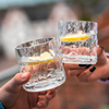 Zwei Hände halten jeweils ein Glas mit klarem Getränk und Zitronenscheiben, unscharfer Hintergrund mit Menschen