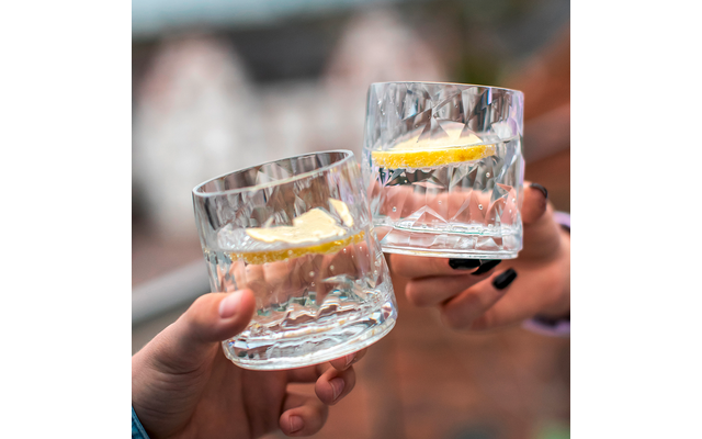 Zwei Hände halten jeweils ein Glas mit klarem Getränk und Zitronenscheiben, unscharfer Hintergrund mit Menschen