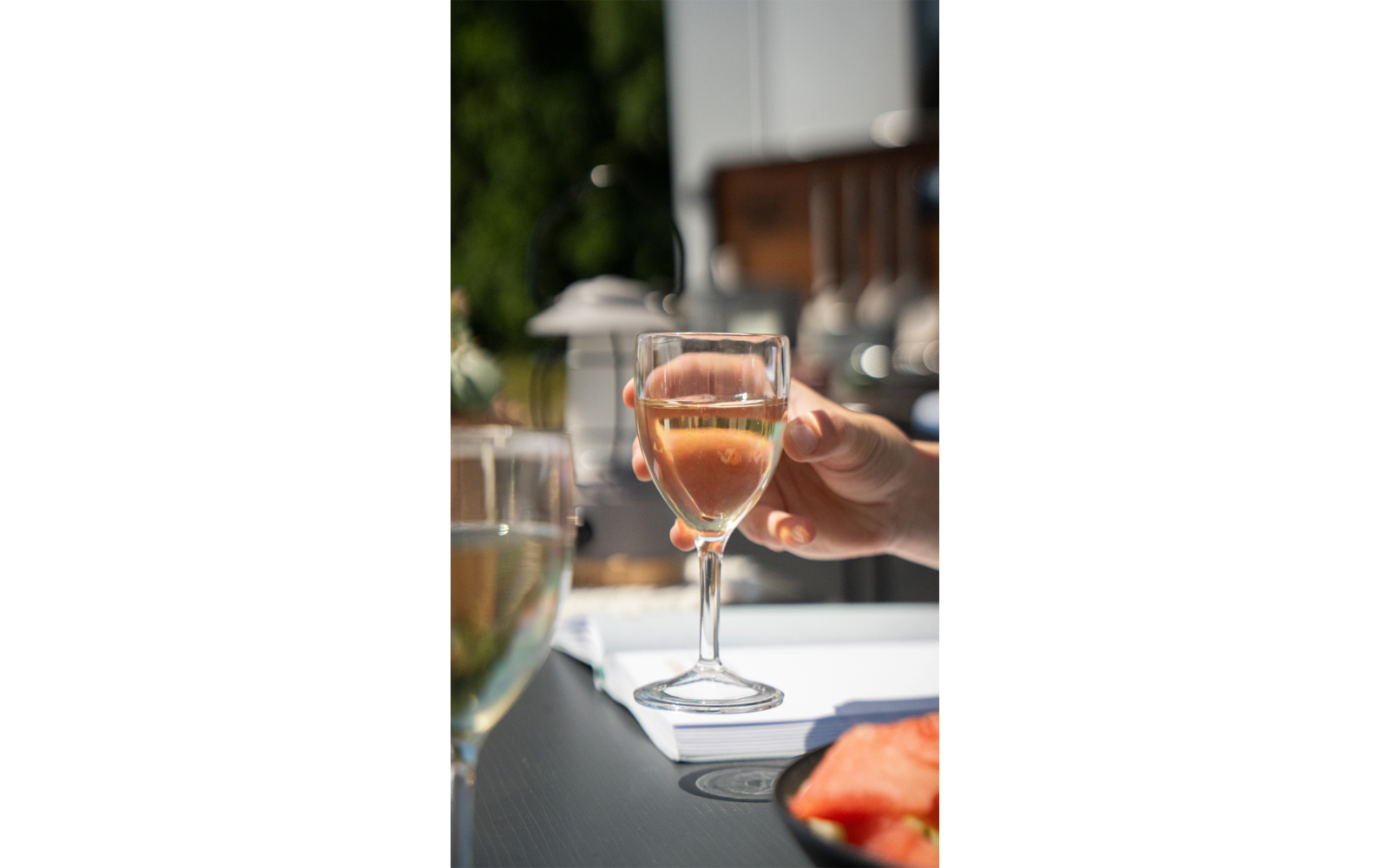 Hand houdt een wijnglas met rosékleurige drank, vage tafel en stoel buiten op de achtergrond