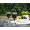 Dos copas de vino tinto sobre una mesa de madera, junto a un cuenco con uvas verdes y oscuras, una planta en maceta y un paño blanco, fondo verde desenfocado
