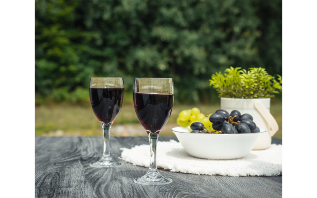 Dos copas de vino tinto sobre una mesa de madera, junto a un cuenco con uvas verdes y oscuras, una planta en maceta y un paño blanco, fondo verde desenfocado