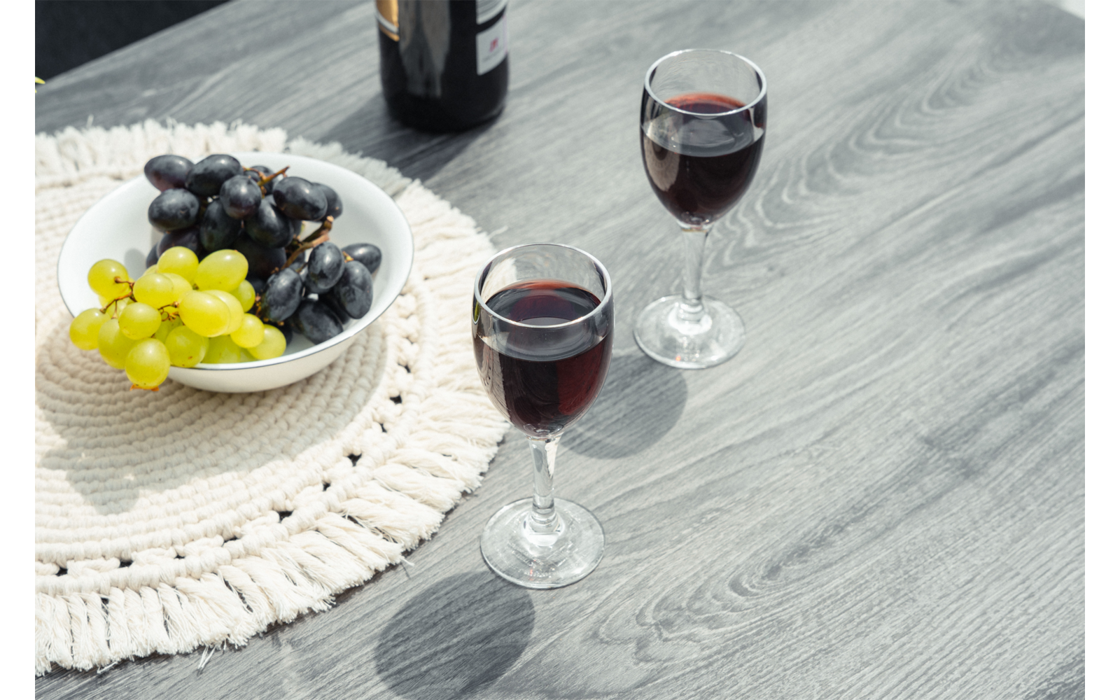 Dos copas de vino tinto en una mesa de madera; junto a ellas un cuenco con uvas verdes y negras sobre un salvamanteles de ganchillo y una botella de vino en el borde