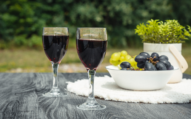 Zwei Weingläser mit Rotwein auf einem Holztisch, daneben eine Schale mit grünen und dunklen Trauben, eine Topfpflanze und ein weißes Tuch, unscharfer grüner Hintergrund