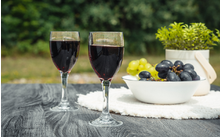 Zwei Weingläser mit Rotwein auf einem Holztisch, daneben eine Schale mit grünen und dunklen Trauben, eine Topfpflanze und ein weißes Tuch, unscharfer grüner Hintergrund