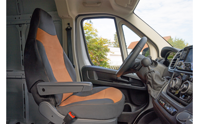 Espace conducteur avec siège gris et orange-brun, accoudoir, volant, tableau de bord et fenêtre latérale montrant des arbres et une maison dehors