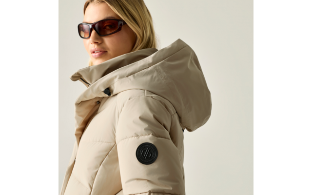 Person mit Sonnenbrille, beige Steppjacke mit Kapuze und rundem Aufnäher auf dem Ärmel