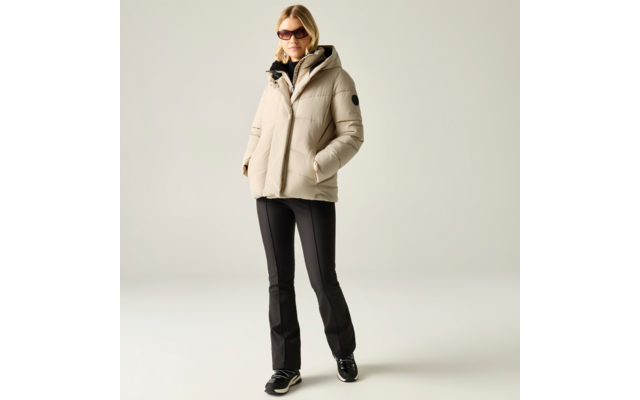 Person vor neutralem Hintergrund, trägt beige Steppjacke mit Kapuze, schwarze ausgestellte Hose, schwarze Sneaker mit weißer Sohle und Sonnenbrille; Hände in den Taschen