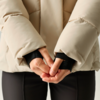 Oberkörper bis Hüfte einer Person, beige gesteppte Jacke, schwarze Innenbündchen, verschränkte Hände vor dem Körper, schwarze Hose