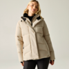 Person trägt beige gesteppte Jacke mit Kapuze und schwarzen Rollkragenpullover, schwarze Hose, steht vor hellgrauem Hintergrund