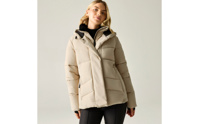Person trägt beige gesteppte Jacke mit Kapuze und schwarzen Rollkragenpullover, schwarze Hose, steht vor hellgrauem Hintergrund