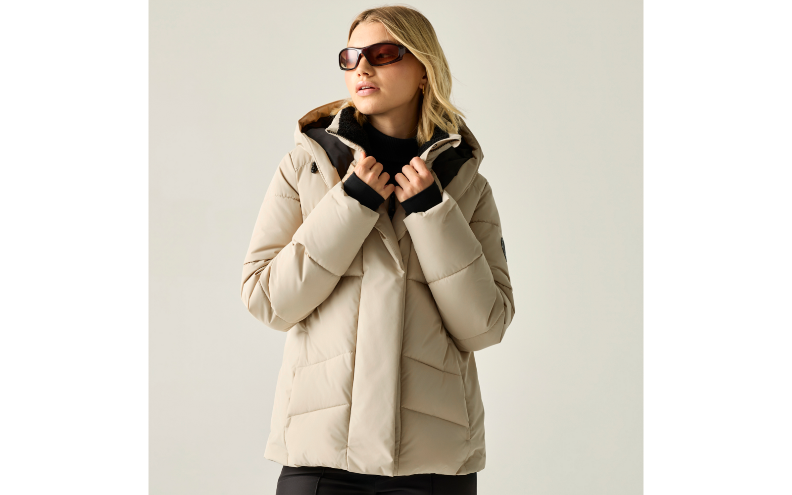 Person mit blonden Haaren trägt beige Steppjacke mit Kapuze und dunkle Sonnenbrille, hält den Jackenkragen mit beiden Händen, vor hellgrauem Hintergrund