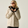 Person mit blonden Haaren trägt beige Steppjacke mit Kapuze und dunkle Sonnenbrille, hält den Jackenkragen mit beiden Händen, vor hellgrauem Hintergrund