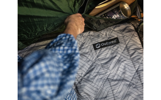 Hand in blau kariertem Hemdsärmel öffnet Reißverschluss eines grünen Schlafsacks, hellgraues Innenfutter mit Linienmuster und Aufnäher 'Outwell', goldfarbene Tasse daneben