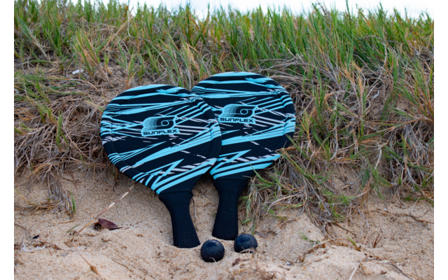 Zwei schwarze Füße ragen unter einem blauen und schwarzen gestreiften Handtuch hervor, im Hintergrund Gras und Sand