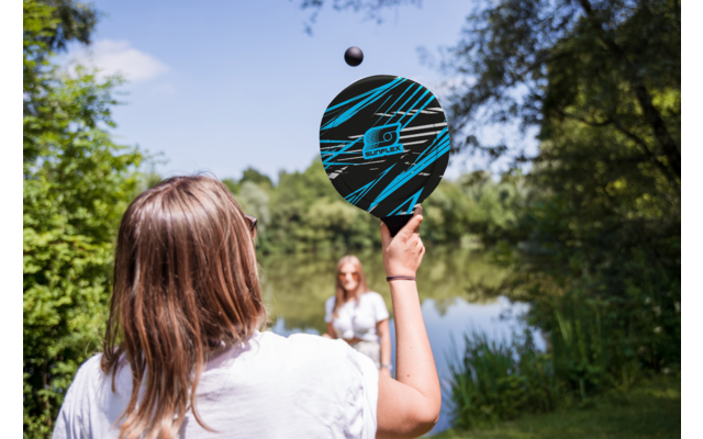 Eine Frau hält einen Basketball mit einer Hand nach oben, im Hintergrund sind Bäume und Himmel sichtbar