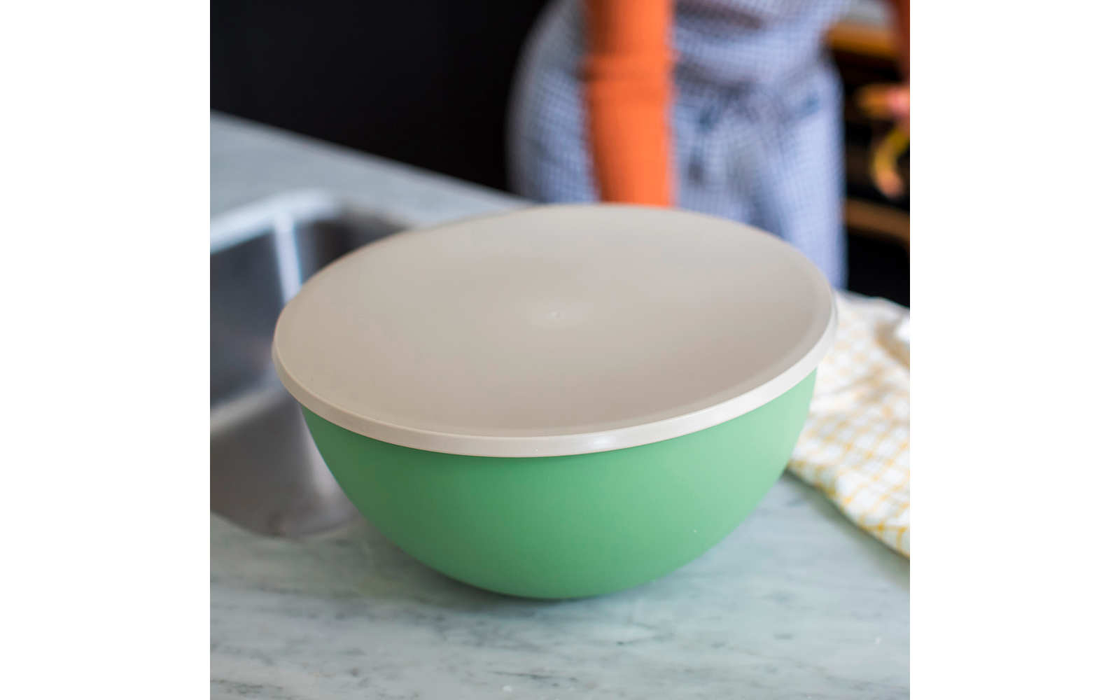 Groene mengkom met wit deksel op marmeren aanrecht, links de gootsteen, rechts een geel theedoek, op de achtergrond een onscherpe persoon met oranje mouw