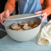 Handen tillen het deksel van een grijze kom met sneetjes stokbrood op een marmeren aanrecht; rechts opgevouwen geel geruit theedoek; persoon draagt oranje trui en blauw geruit schort