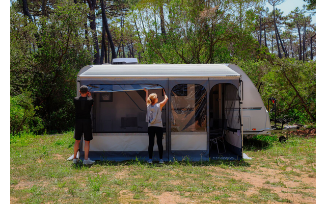 Wohnwagen mit angebautem Vorzelt auf einer Waldlichtung; zwei Personen von hinten, die am Vorzelt arbeiten