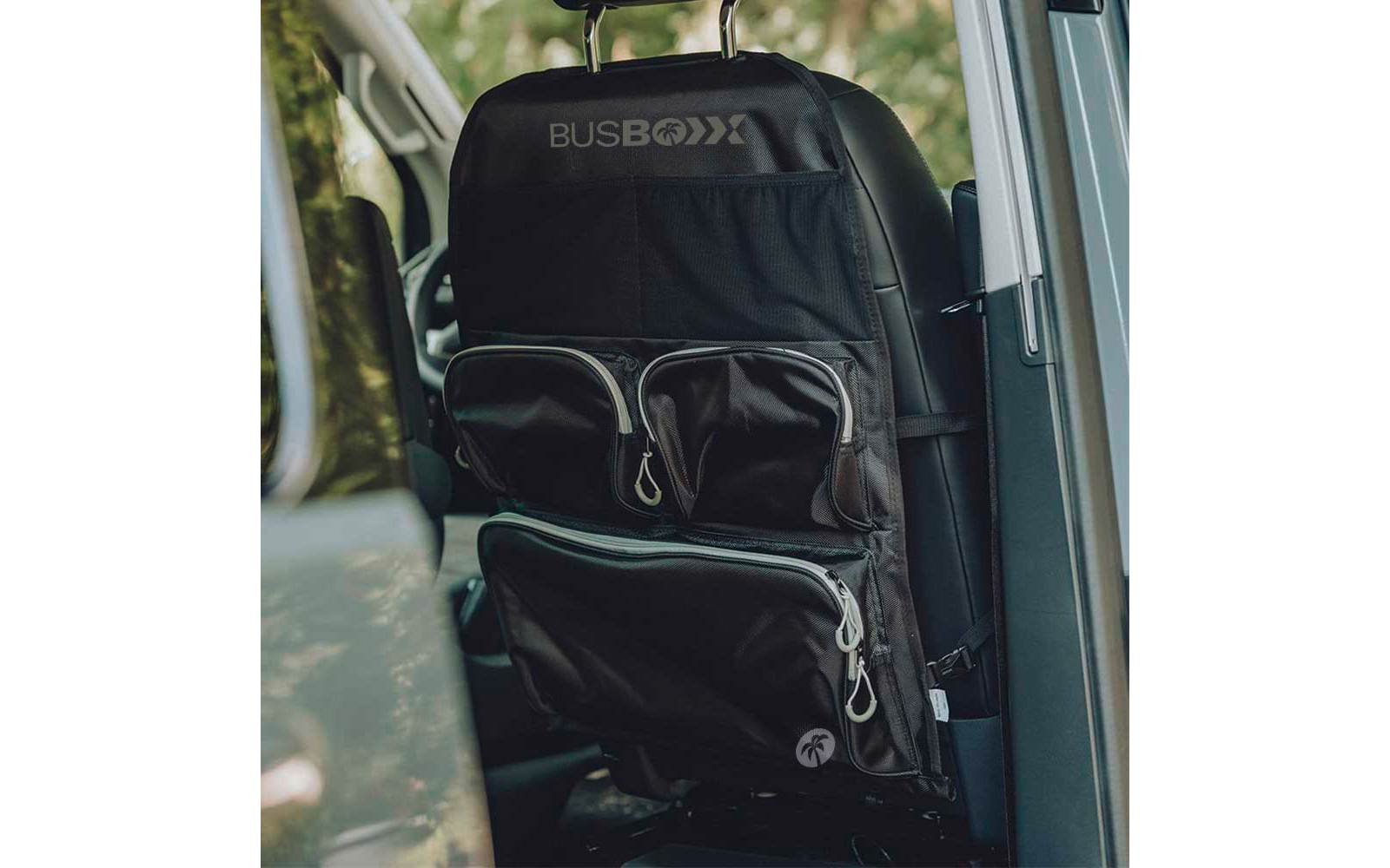 Schwarze Stoff-Organizer-Tasche mit dem Schriftzug "BUSBOXX" an der Rückseite eines Fahrzeugsitzes, mehrere Reißverschlusstaschen, Kopfstützenriemen und Autofenster mit grüner Außenansicht