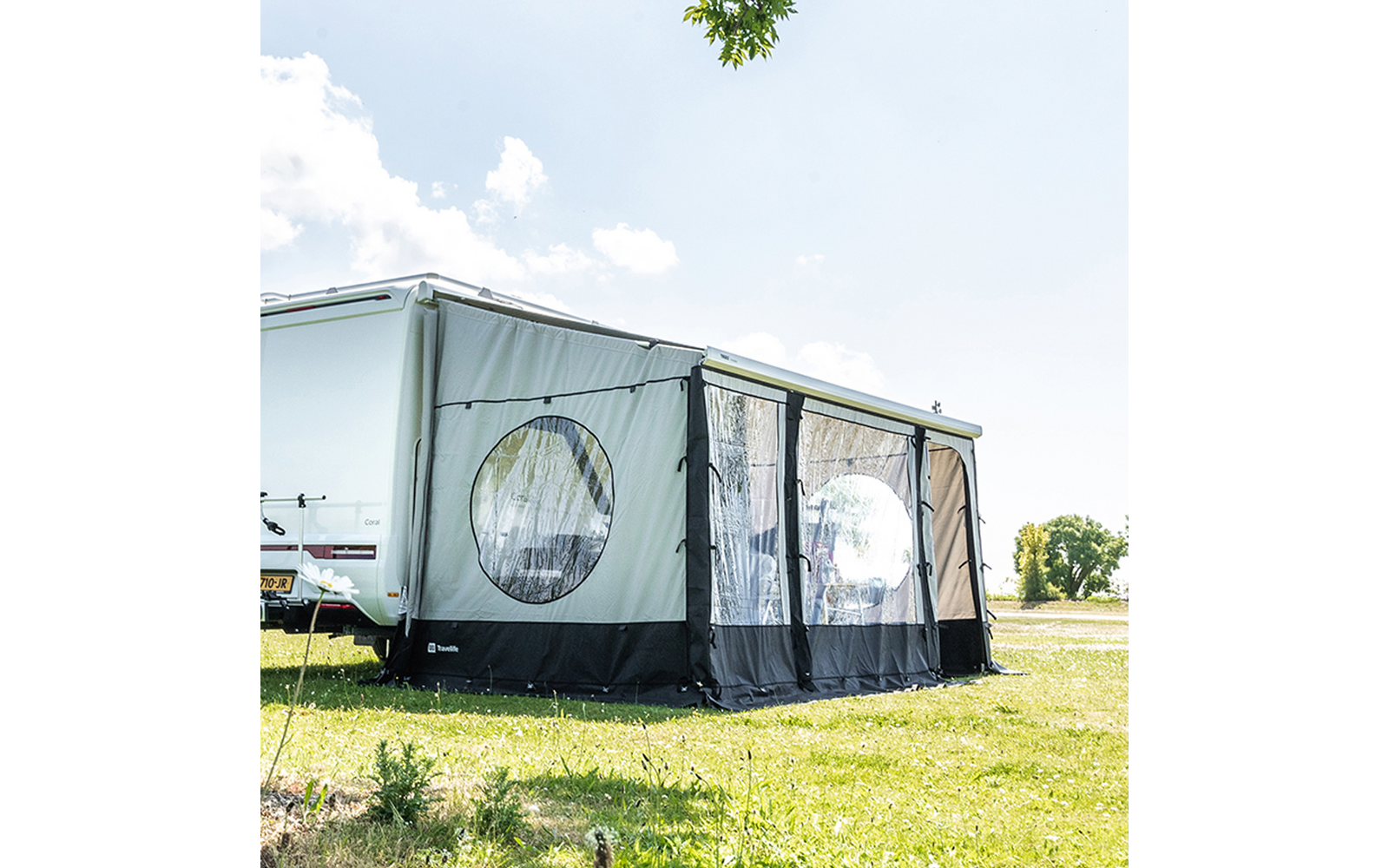 Weißer Wohnwagen mit grauem Vorzelt mit runden und rechteckigen Fenstern auf einer Wiese unter blauem Himmel