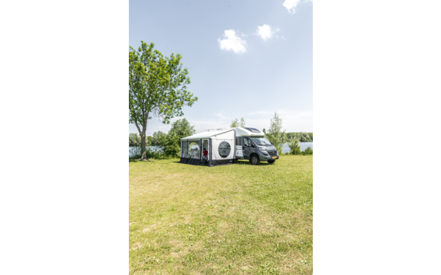 Wohnmobil mit angebautem Vorzelt auf einer Wiese neben einem See, Bäume und blauer Himmel mit vereinzelten Wolken