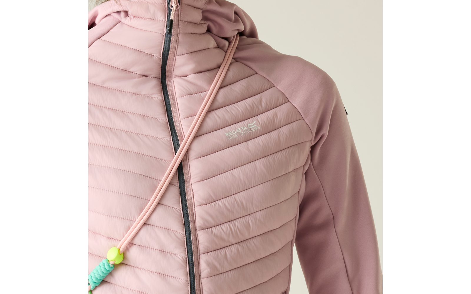 Nahaufnahme des Vorderteils einer rosafarbenen, gestepp­ten Kapuzenjacke mit vertikalem Reißverschluss, Kordel mit grünem Stopper und kleinem weißen Logo auf der Brust