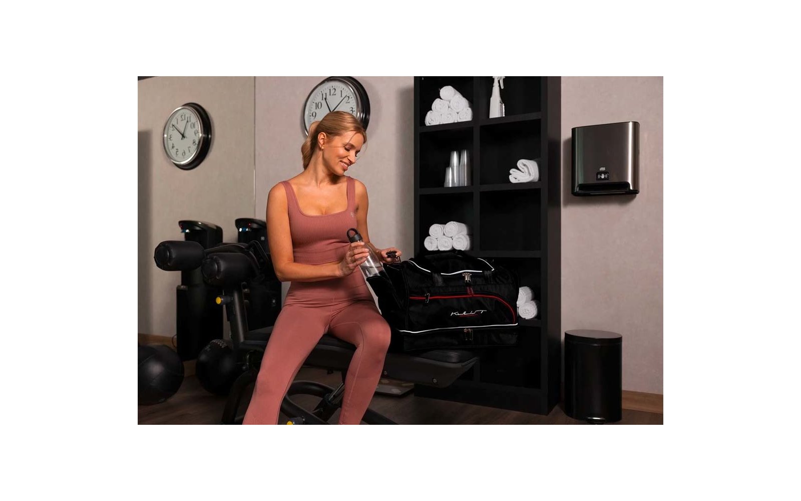 Femme en tenue de sport rose assise sur un banc, tenant une bouteille à côté d'un sac de sport noir ouvert; étagère avec serviettes blanches, horloge murale et appareils de fitness en arrière-plan