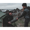 Zwei Männer in Outdoor-Kleidung beim Umgang mit einem Zeltausleger am Meer mit Felsen im Hintergrund