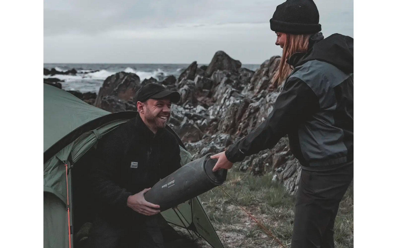 Zwei Männer in Outdoor-Kleidung beim Umgang mit einem Zeltausleger am Meer mit Felsen im Hintergrund