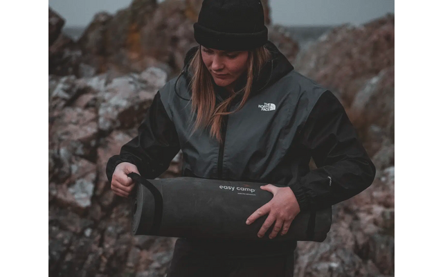 Frau in Outdoor-Bekleidung hält eine zusammengerollte Matte und steht vor felsigem Hintergrund