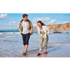 Un homme et une femme souriants marchent dans l'eau peu profonde sur une plage; tous deux portent des sandales; falaises rocheuses et ciel nuageux en arrière-plan