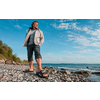 Homme en veste claire et pantalon retroussé debout sur une plage de galets, sandales sur les pierres, mer et ciel nuageux en arrière-plan