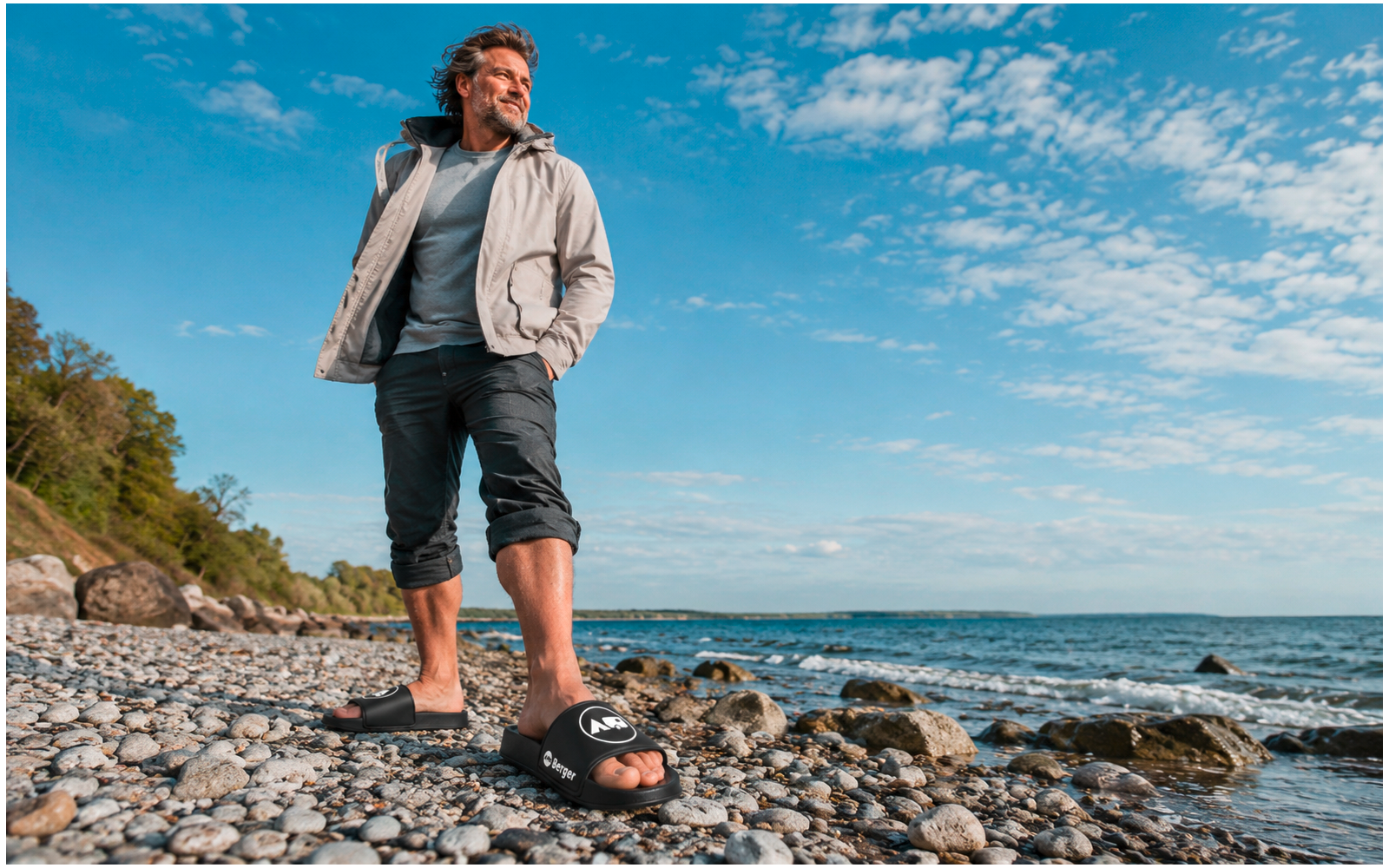 Homme en veste claire et pantalon retroussé debout sur une plage de galets, sandales sur les pierres, mer et ciel nuageux en arrière-plan