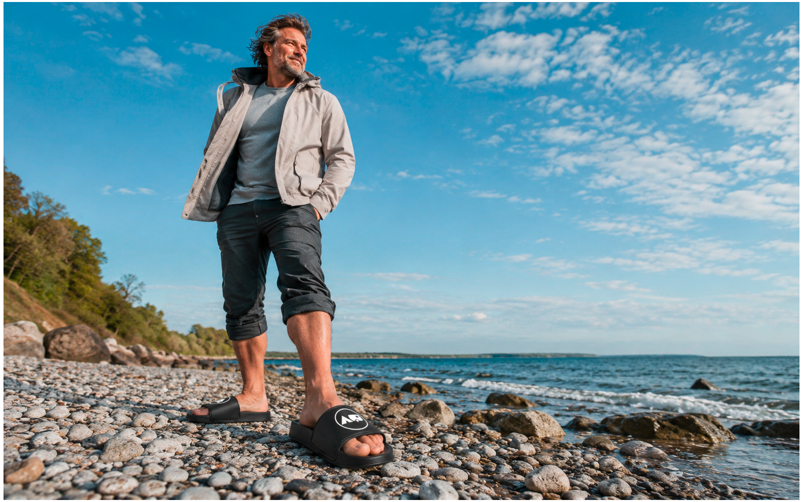 Mann in heller Jacke und hochgekrempelter Hose steht auf Kiesstrand, Sandalen auf den Steinen, Meer und bewölkter Himmel im Hintergrund