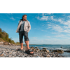 Mann in heller Jacke und hochgekrempelter Hose steht auf Kiesstrand, Sandalen auf den Steinen, Meer und bewölkter Himmel im Hintergrund