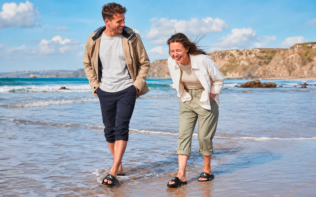 Un homme et une femme souriants marchent dans l'eau peu profonde sur une plage; tous deux portent des sandales; falaises rocheuses et ciel nuageux en arrière-plan