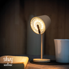 Eingeschaltete Tischlampe, daneben aufgeschlagenes Buch, Tasse auf dunklem Untersetzer, holzvertäfelte Wand im Hintergrund, Markenlogo 'silvy x sompex' unten links