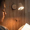 Unscharfe Person im Vordergrund links, Hand hält aufgeschlagenes Buch rechts, eingeschaltete Wandlampe an Holzverkleidung