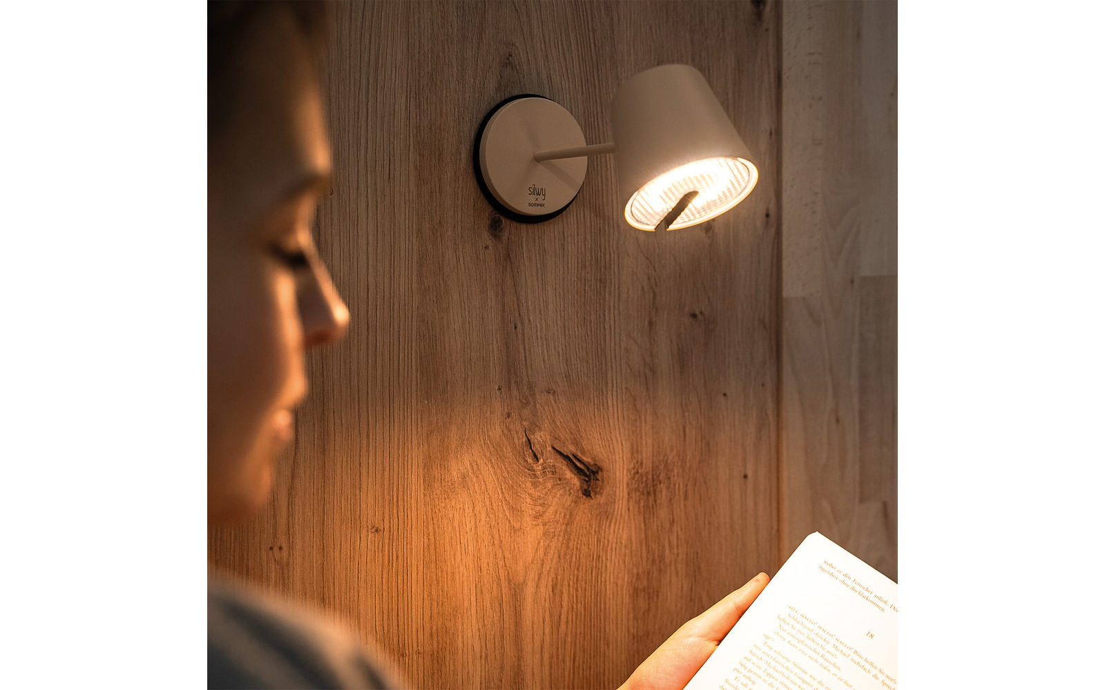 Unscharfe Person im Vordergrund links, Hand hält aufgeschlagenes Buch rechts, eingeschaltete Wandlampe an Holzverkleidung